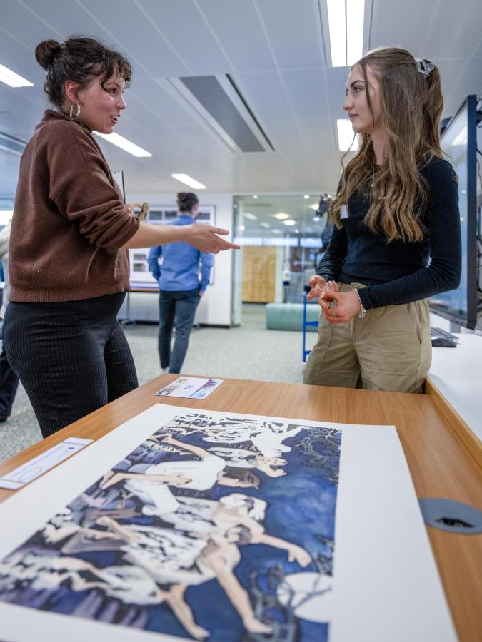 In the foreground a large artwork shows ballet dancers performing. In the background two people are in conversation about the artwork.